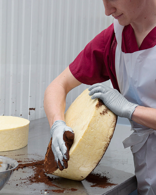 A gloved hand rubbing coffee on a 20 pound wheel of cheddar